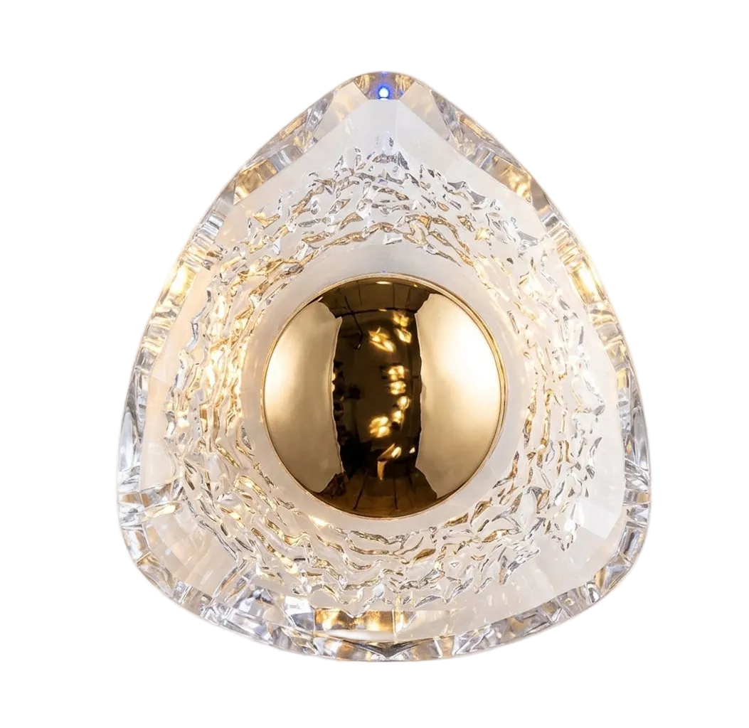 A modern geometric wall sconce featuring a clear faceted triangular crystal-like shade with a textured inner ring and a central gold metallic sphere, illuminated from within against a plain light background.